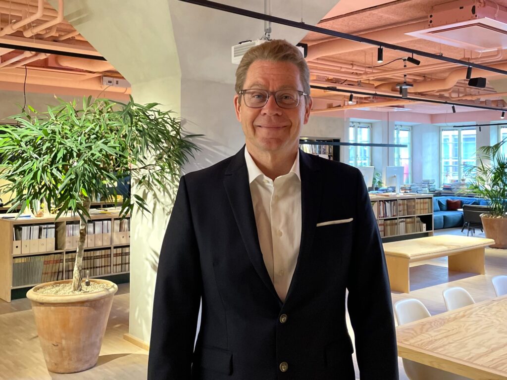En man i kostym och glasögon står och ler i ett kontor med växter, bokhyllor och stora fönster i bakgrunden.