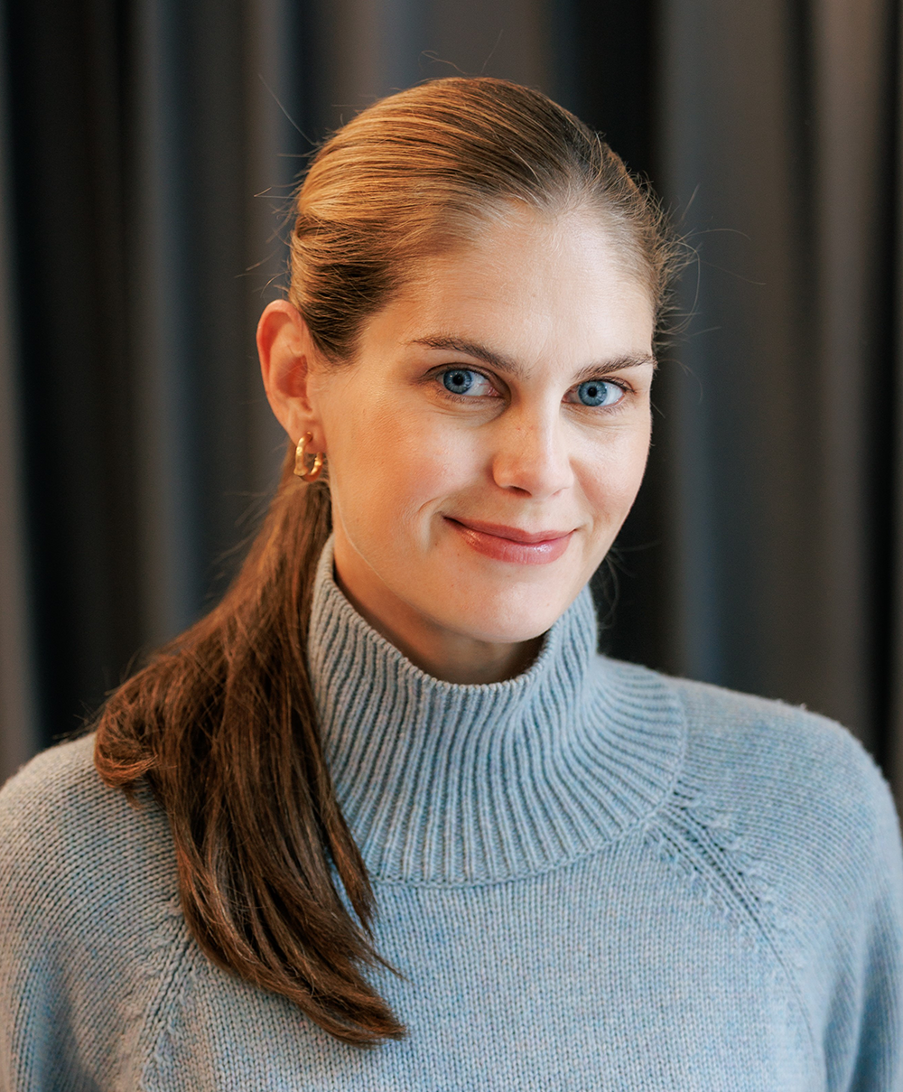 En kvinna med långt brunt hår i hästsvans och ljusblå polotröja står framför mörka gardiner och ler svagt mot kameran.