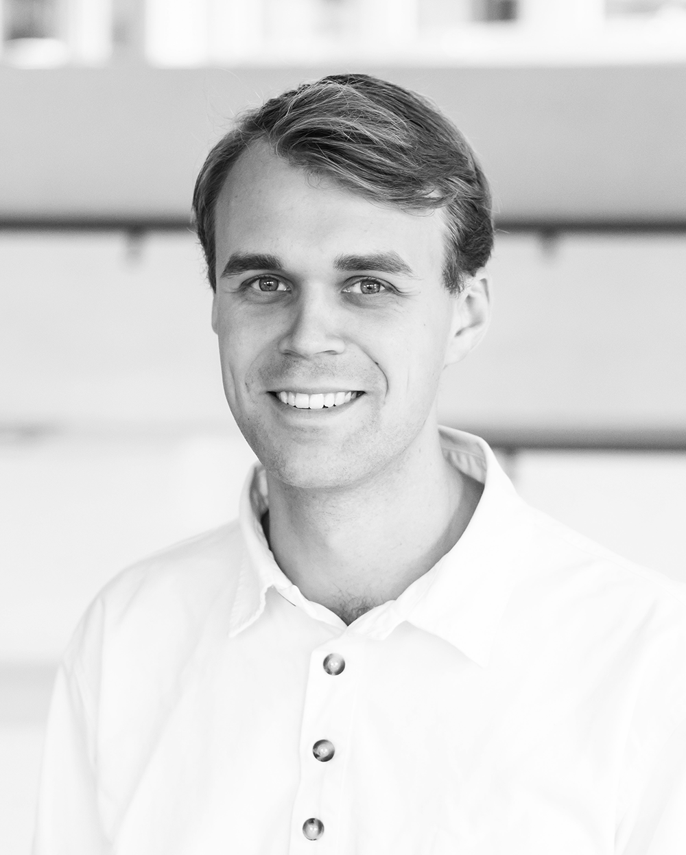 En man med ljust hår och vit skjorta ler mot kameran i en inomhusmiljö.