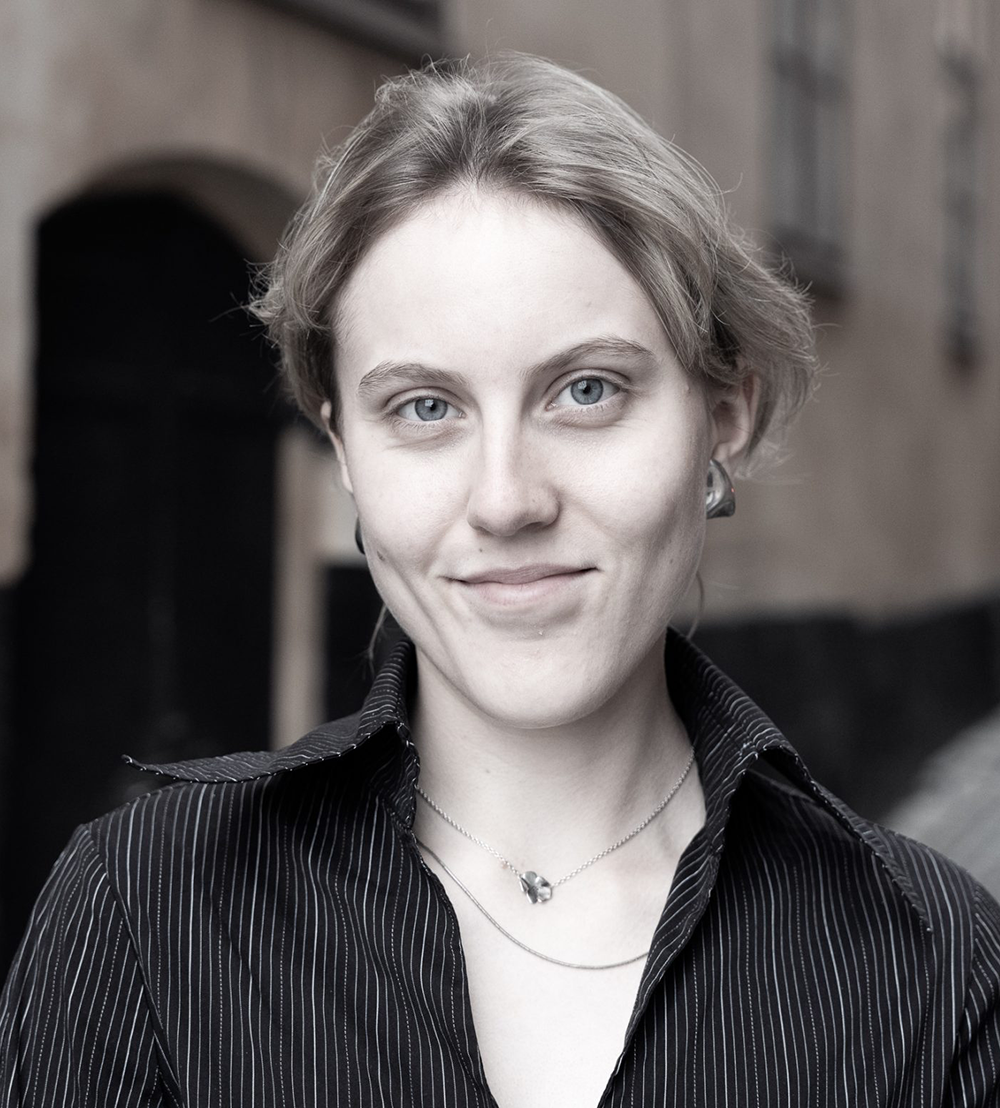 En ung person med ljust hår och ljusa ögon har på sig en mörk skjorta med ränder och ett halsband, utomhus framför en suddig byggnad.