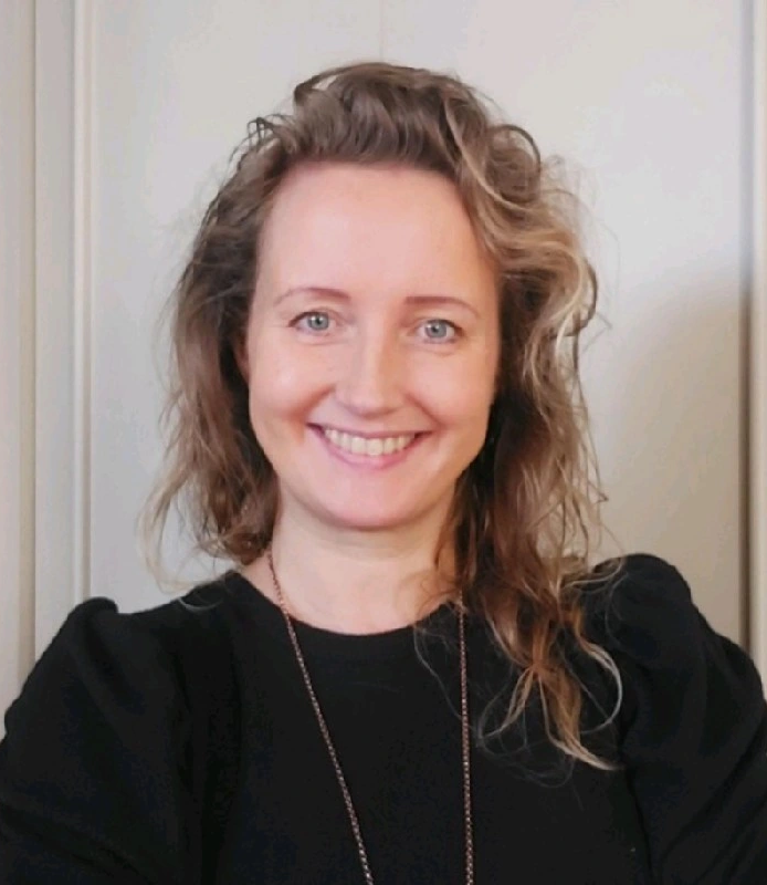 En kvinna med vågigt, axellångt hår och en svart topp ler mot kameran framför en neutral bakgrund.