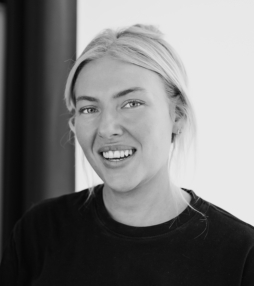 En kvinna med ljust hår i en hästsvans och mörk tröja ler mot kameran på detta svartvita porträtt.