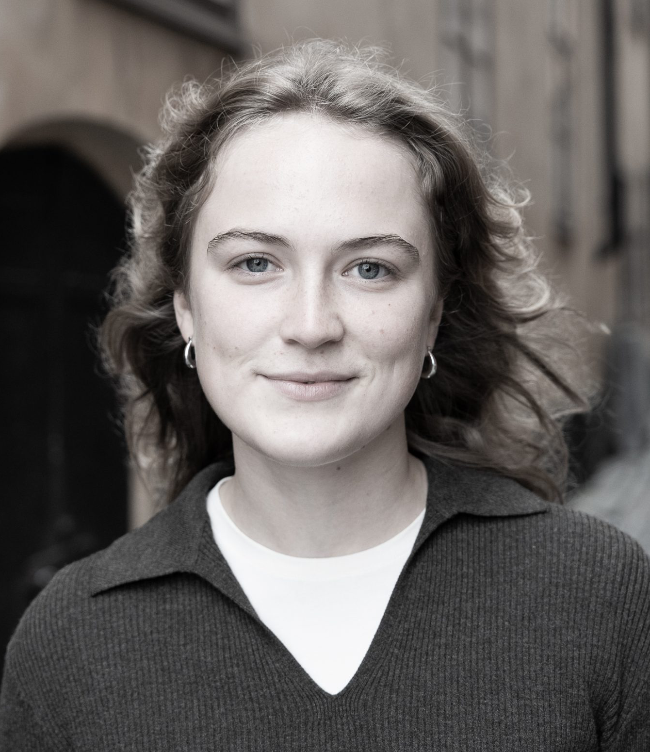 En ung person med vågigt hår, som har runda örhängen och en mörk skjorta över en vit tröja, står ute och tittar mot kameran.