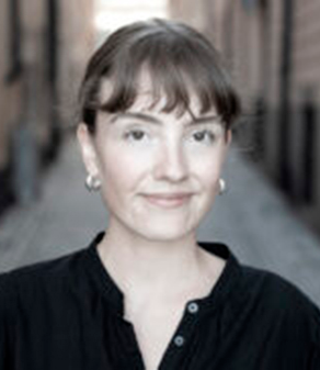 En person med ljus hud, kort brunt hår med lugg och ringörhängen, har en svart topp och står utomhus på en smal gata.