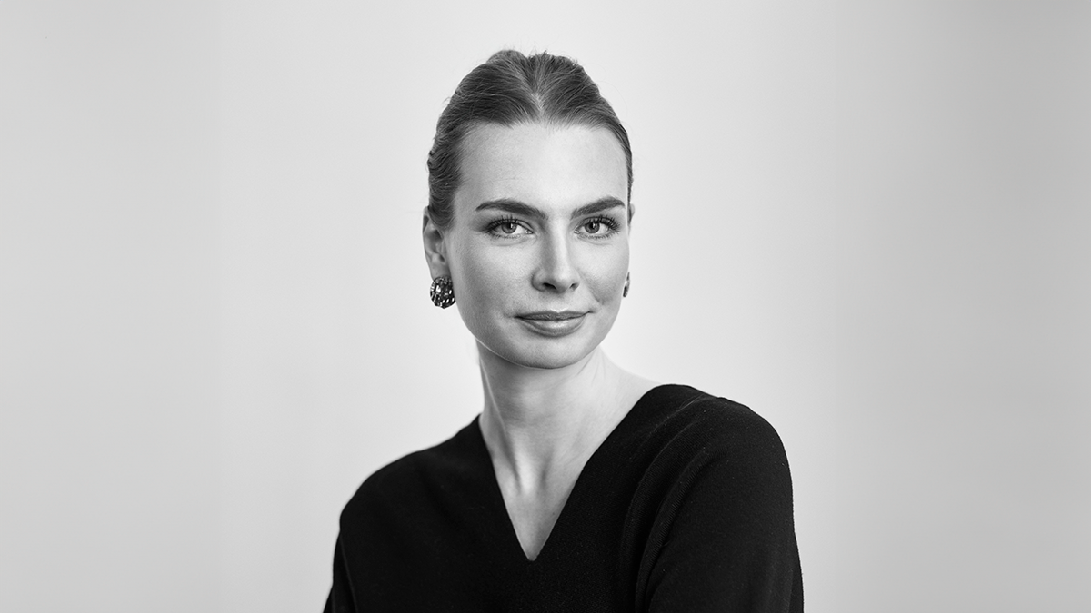 En kvinna med bakåtdraget hår, i mörk topp och örhängen, tittar mot kameran framför en ljus bakgrund.