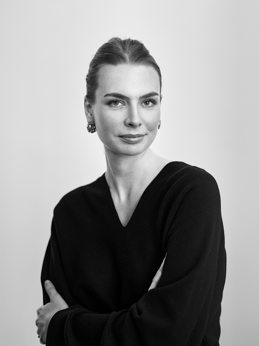 En kvinna med bakåtdraget hår, i mörk topp och örhängen, tittar mot kameran framför en ljus bakgrund.