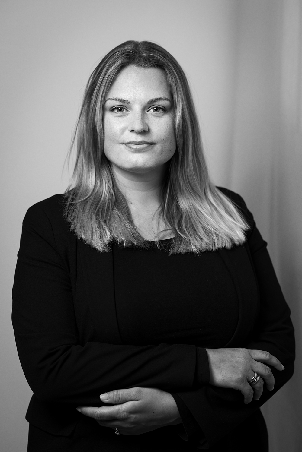 En kvinna med långt, ljust hår har på sig en mörk kavaj och topp och står med armarna i kors i en neutral miljö.