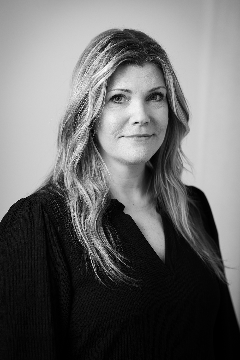 En kvinna med långt, vågigt hår och mörk tröja tittar mot kameran med neutral min i ett svartvitt porträtt.