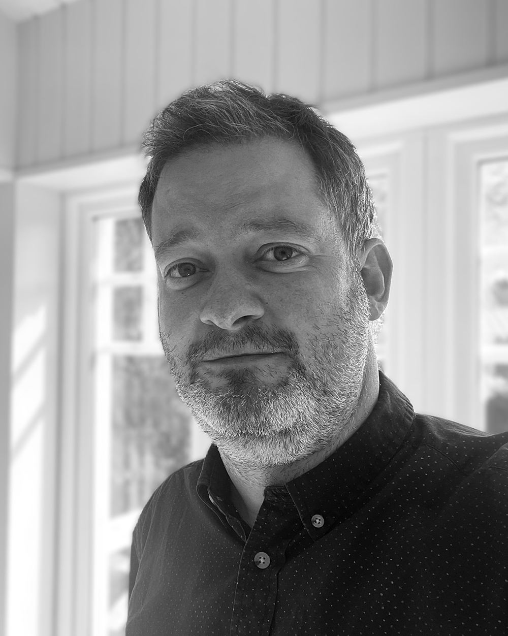 Svartvit porträttbild av en man med kort hår och skägg, i mörk skjorta, står inomhus framför stora fönster.
