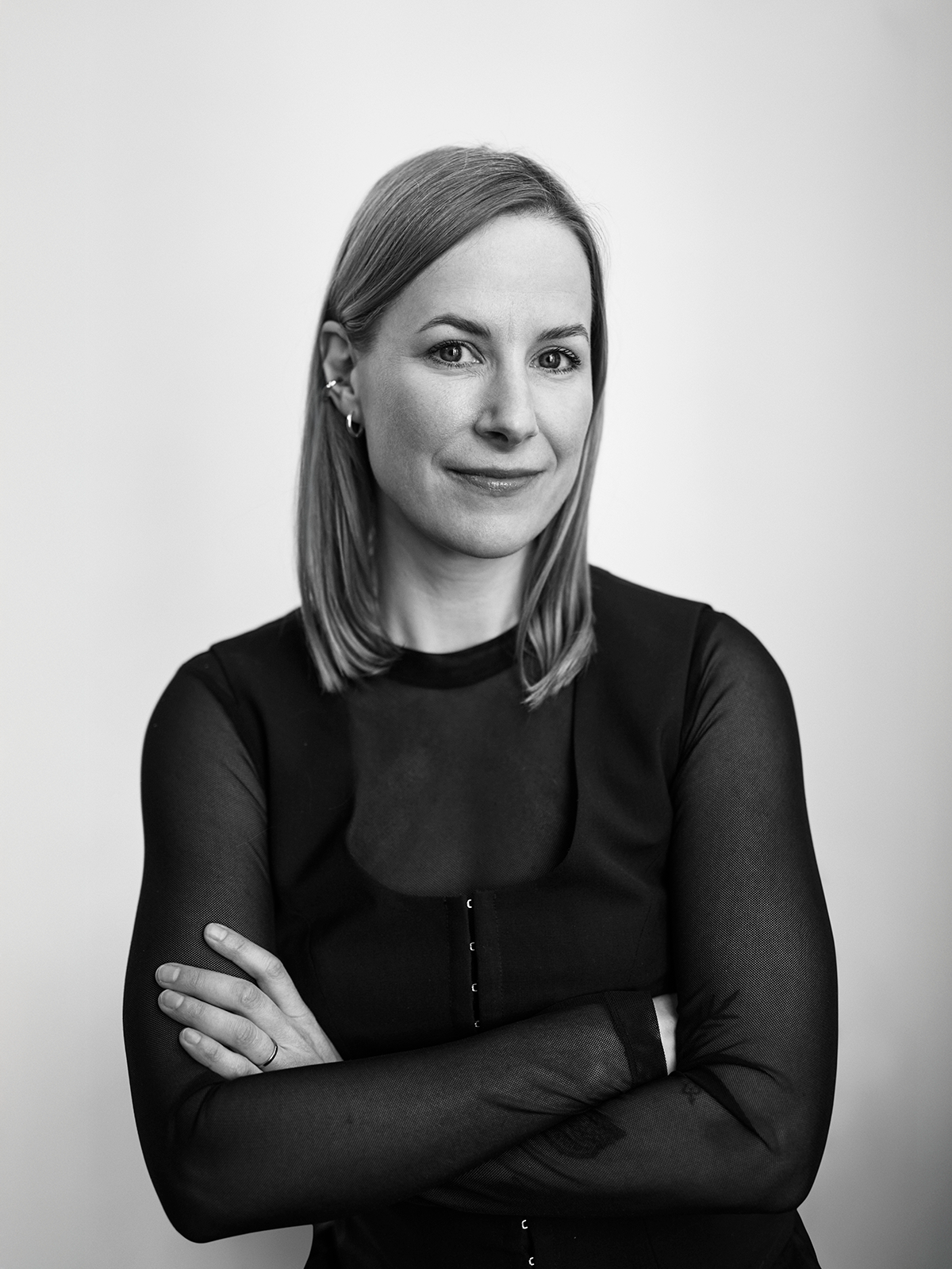 En kvinna med rakt axellångt hår står med armarna i kors, har på sig en topp med genomskinliga ärmar och tittar mot kameran.