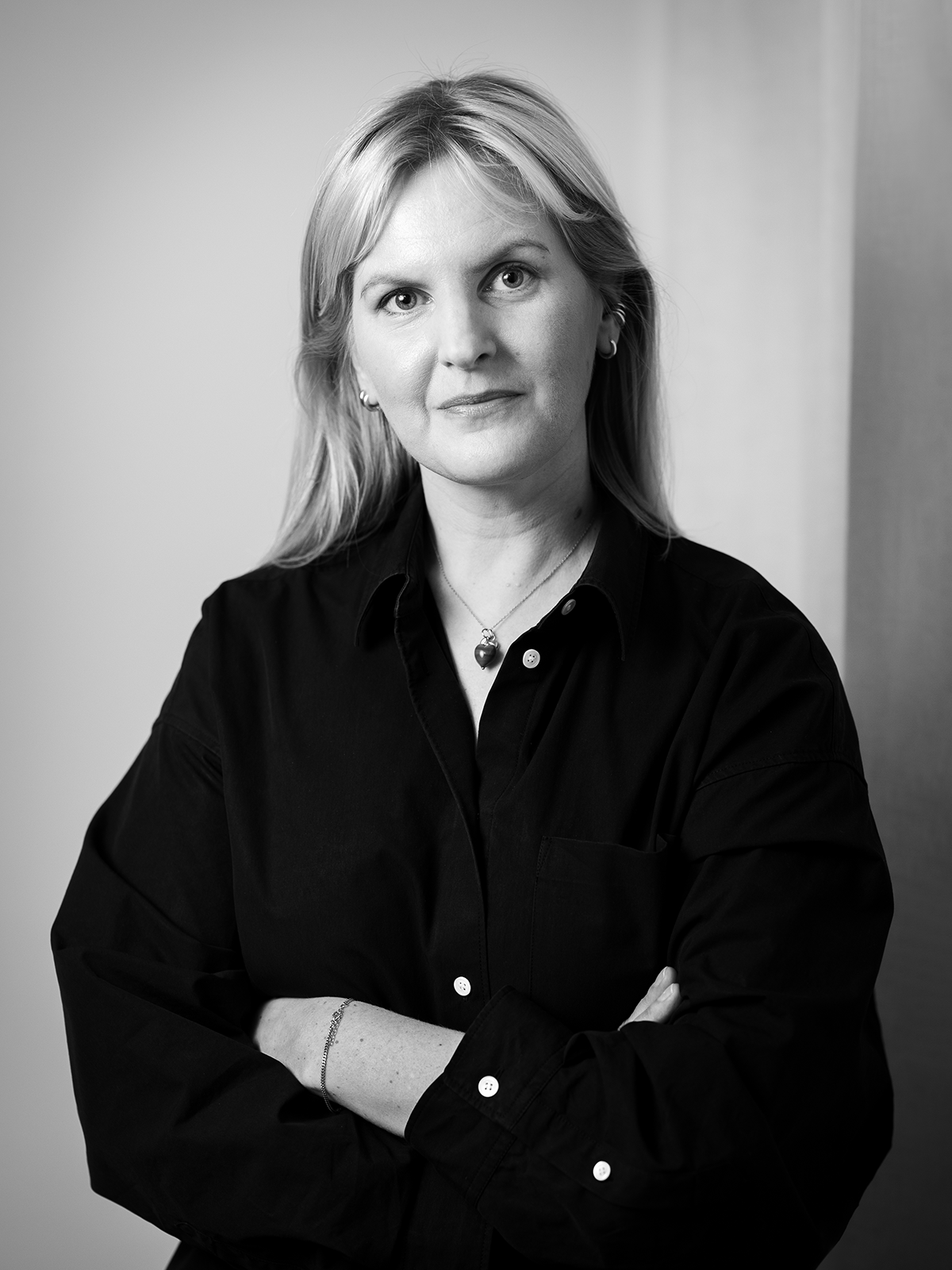 En kvinna med ljust hår bär en mörk skjorta och står med armarna i kors, tittande mot kameran i ett svartvitt porträtt.