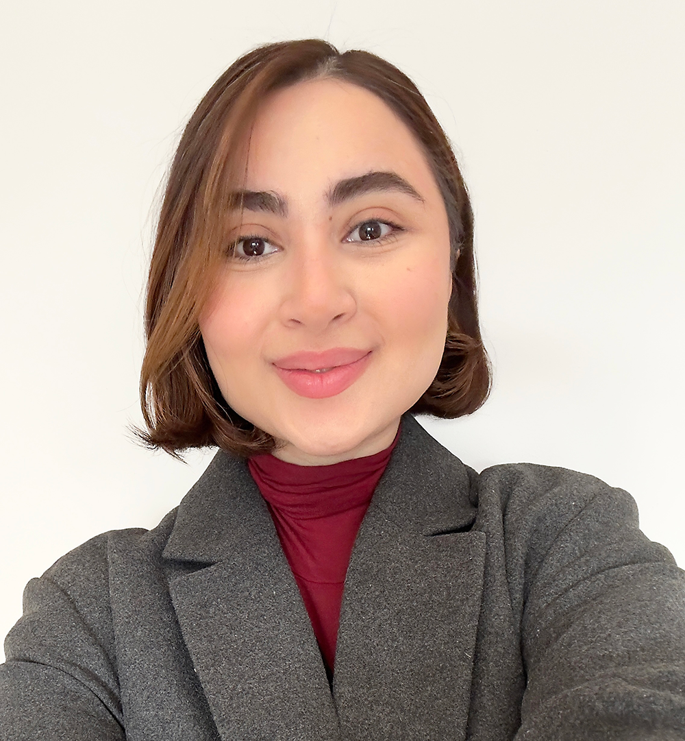 En kvinna med kort brunt hår, grå kavaj och vinröd polotröja ler mot kameran framför en vit bakgrund.