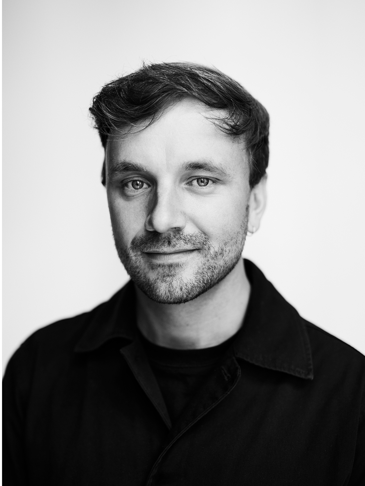 Porträtt i svartvitt av en man med kort hår och ljus skäggväxt, bär mörk kragskjorta, ser in i kameran mot enkel bakgrund.
