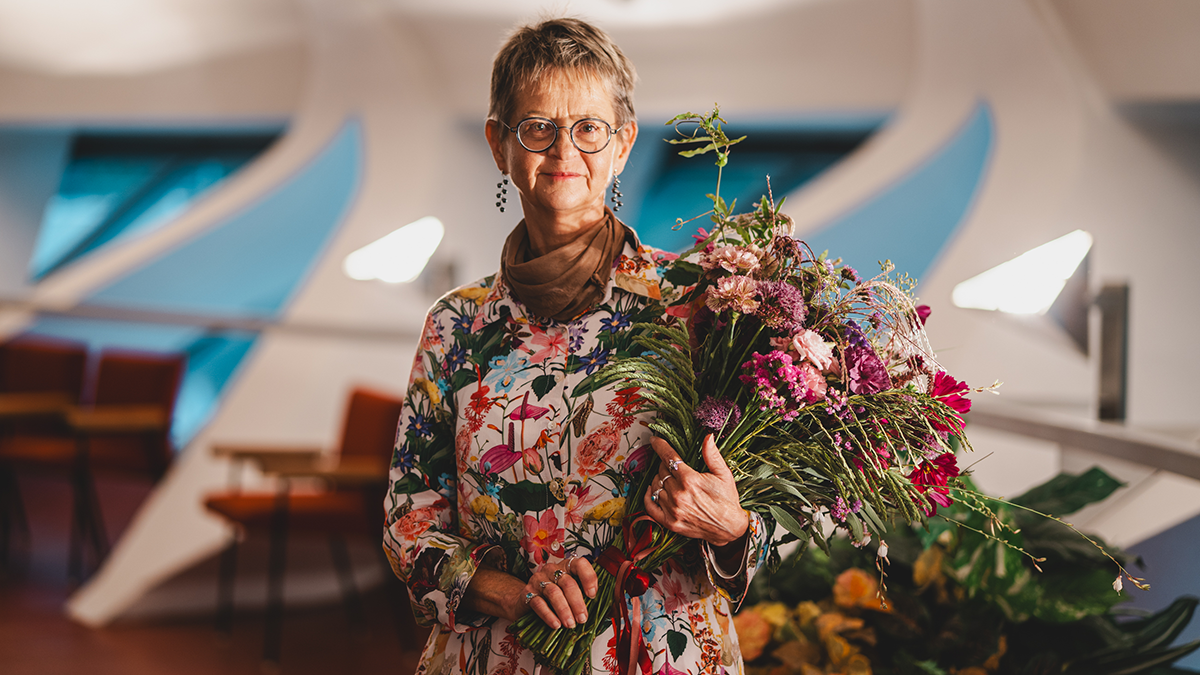 En kvinna med glasögon och blommig klänning står inomhus och håller en bukett blommor, med möbler i bakgrunden.