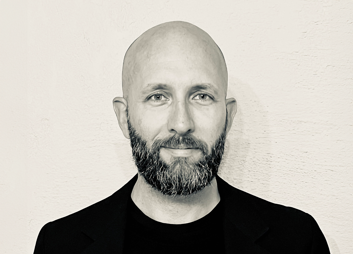 En skallig man med skägg och mustasch står framför en ljus vägg och tittar mot kameran.
