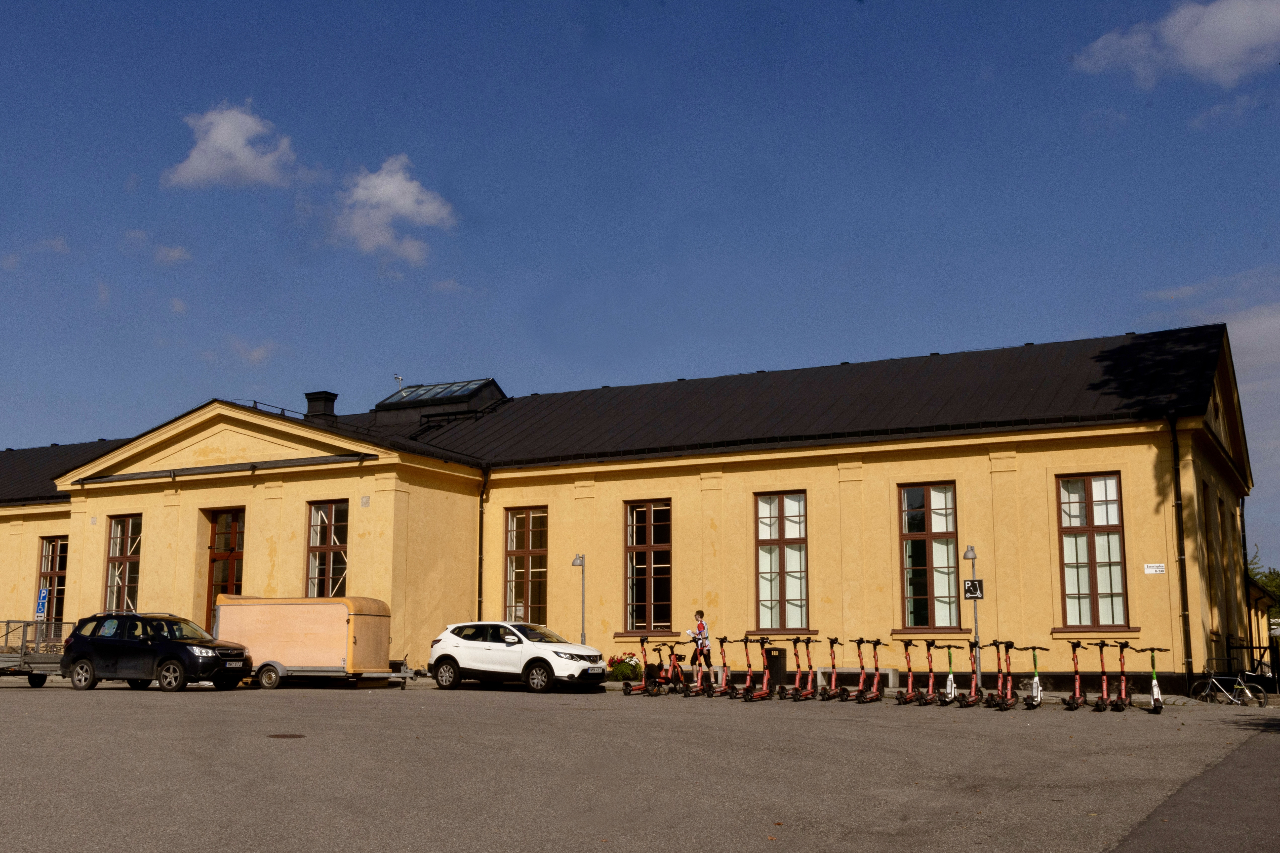 En gul byggnad med stora fönster, flera parkerade bilar och en rad elsparkcyklar utanför under en blå himmel.