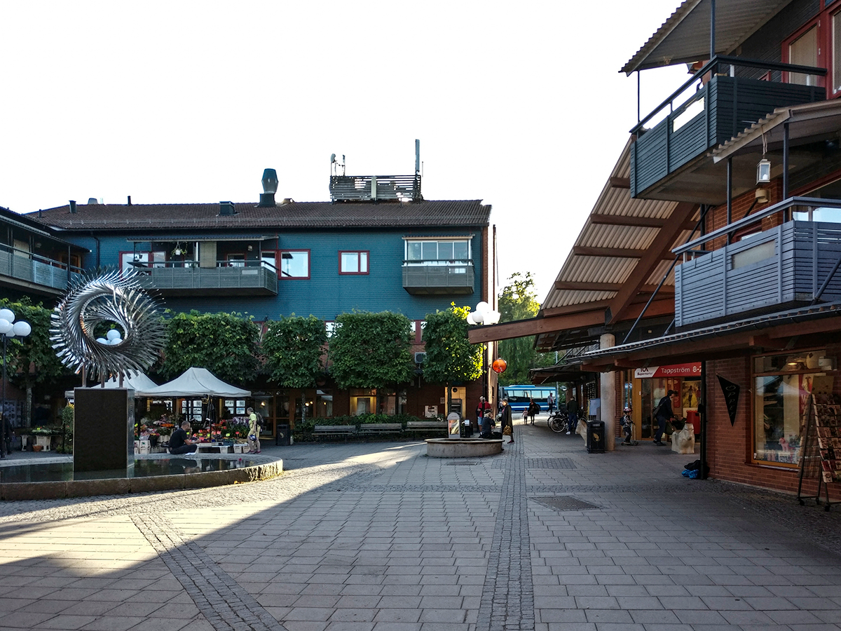 Utomhustorg med byggnader, butiker, några personer som sitter, träd och en metallskulptur på sockel i förgrunden.