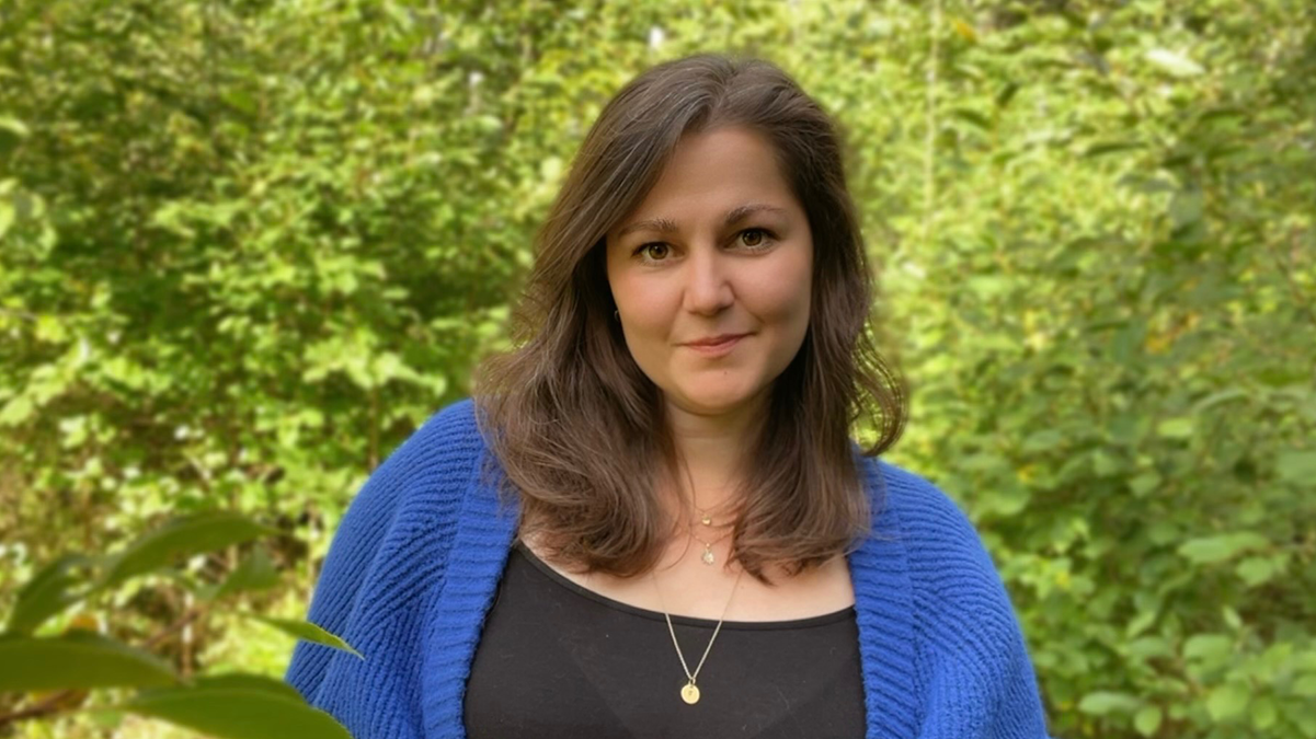 En kvinna med brunt hår, blå tröja och svart topp står utomhus framför grönska.