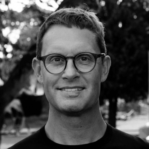 En man med kort hår och glasögon ler mot kameran utomhus, med träd och suddig bakgrund bakom sig.