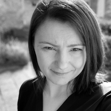 En kvinna med rakt mörkt hår är utomhus och tittar in i kameran med neutral min. Hon har på sig en mörk topp. Bakgrunden är suddig.