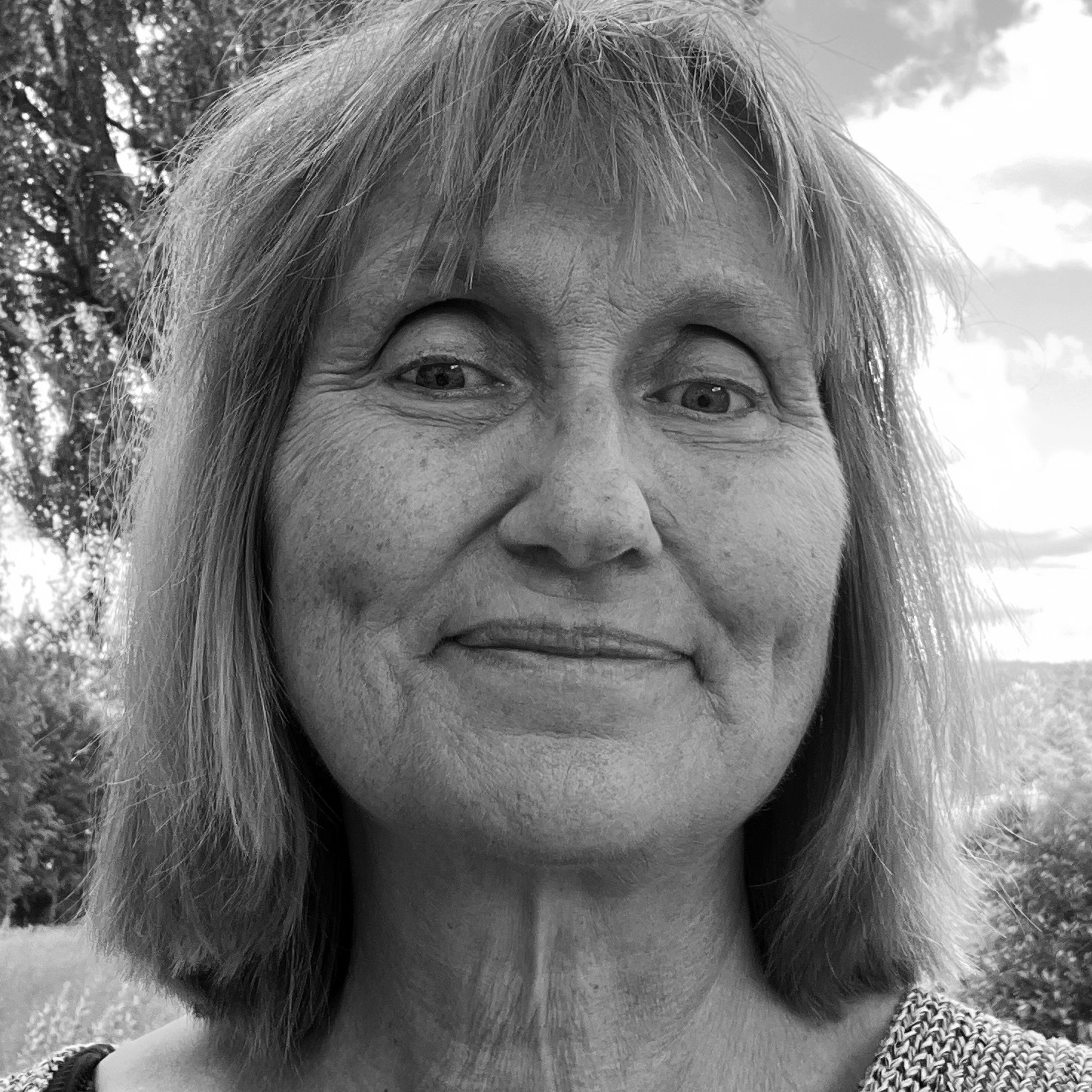 En kvinna med rakt, axellångt hår är utomhus, tittar in i kameran och ler svagt. Bakom henne finns träd och moln. Bilden är svartvit.