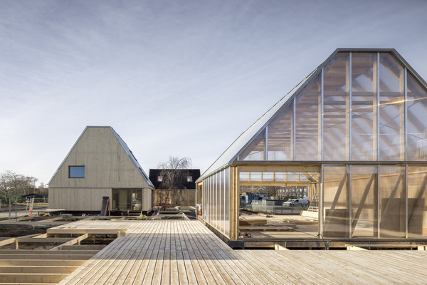 Två byggnader med sadeltak under konstruktion har trä- och genomskinliga paneler, förbundna av en gång i trä en klar dag.