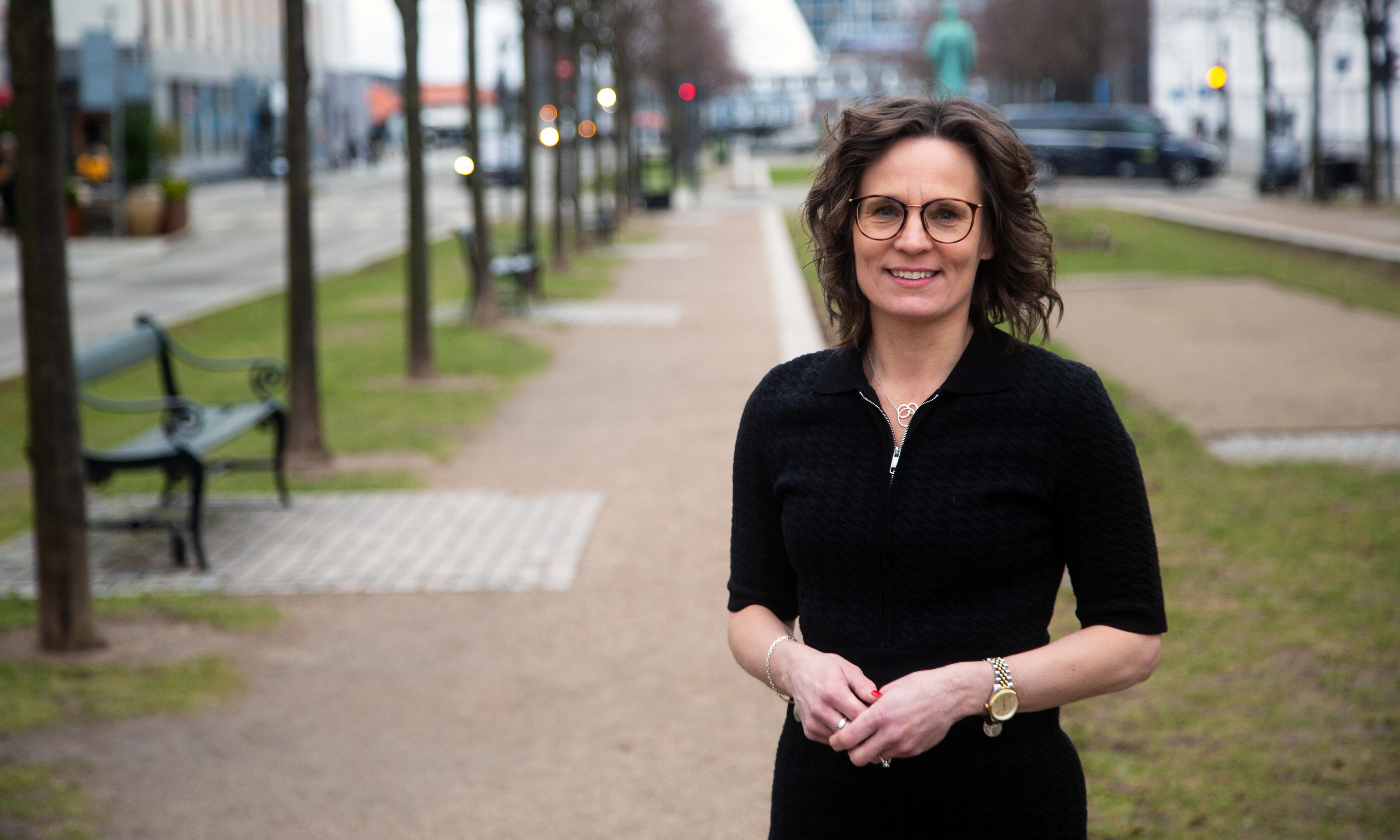 En kvinna med glasögon och mörkt hår står på en gångväg i en stadspark, tittar mot kameran med byggnader och träd bakom sig.
