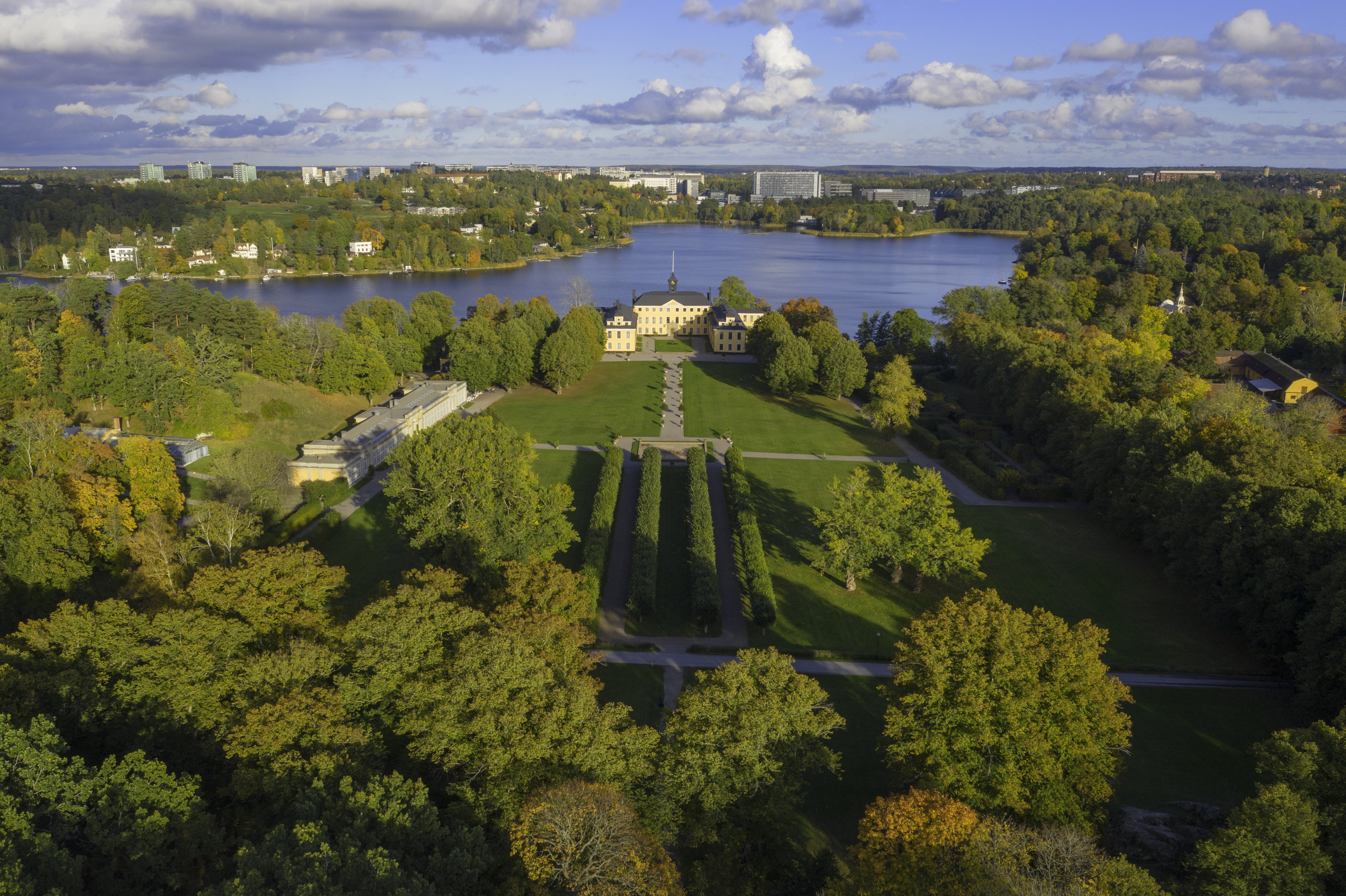 Flygfoto av en stor gul herrgård omgiven av symmetriska trädgårdar, träd med höstlöv och en sjö framför stadsbyggnader.