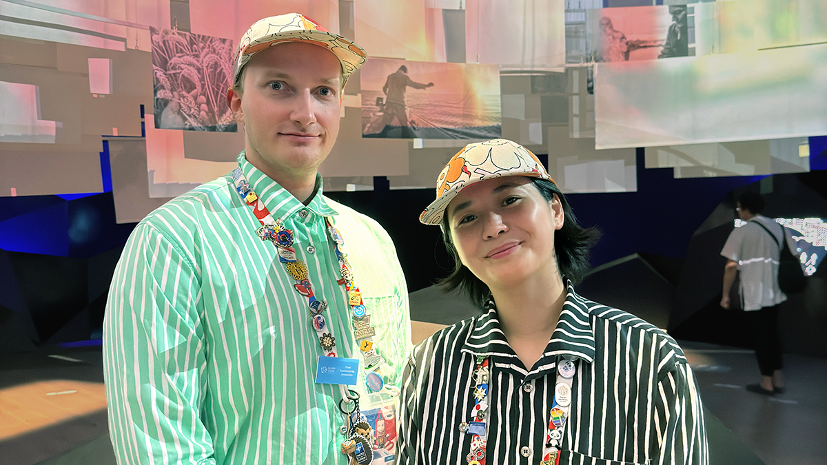 Två personer med randiga tröjor, mönstrade nyckelband och kepsar står inomhus framför en display med hängande skärmar och lampor.