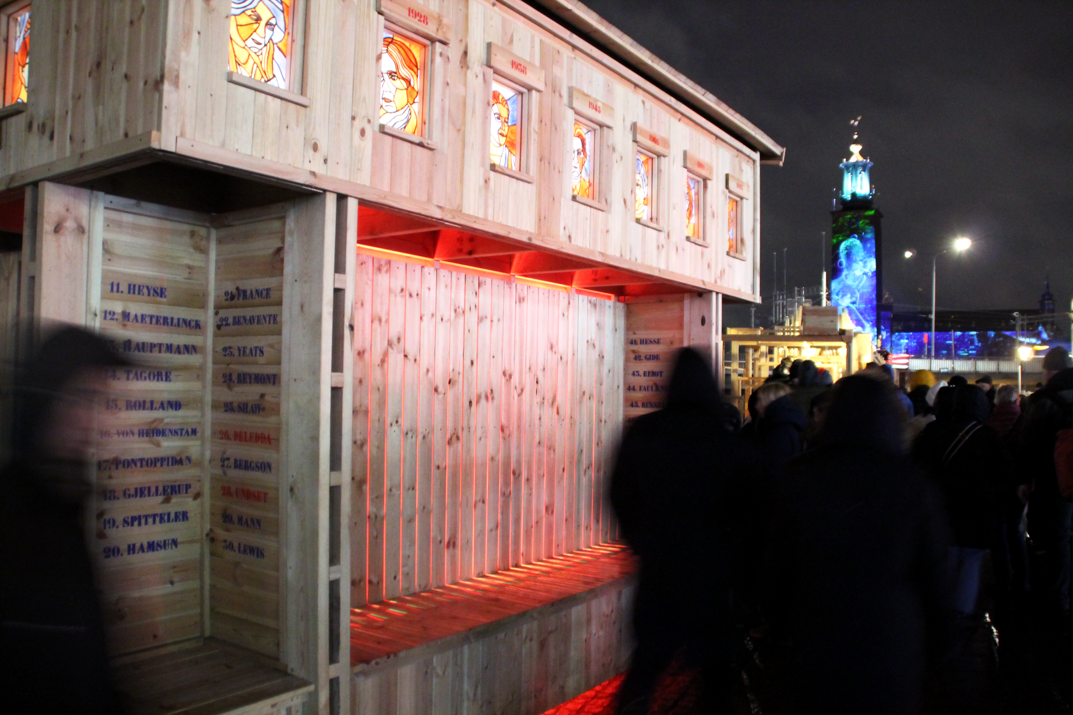 En trästruktur med belysta färgade glasfönster visas på natten, medan människor går bredvid under konstgjort ljus utomhus.