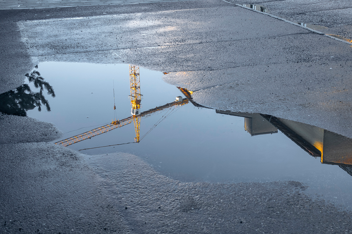 En byggkran och en del av en byggnad speglas i en stor vattenpöl på en asfaltsyta.