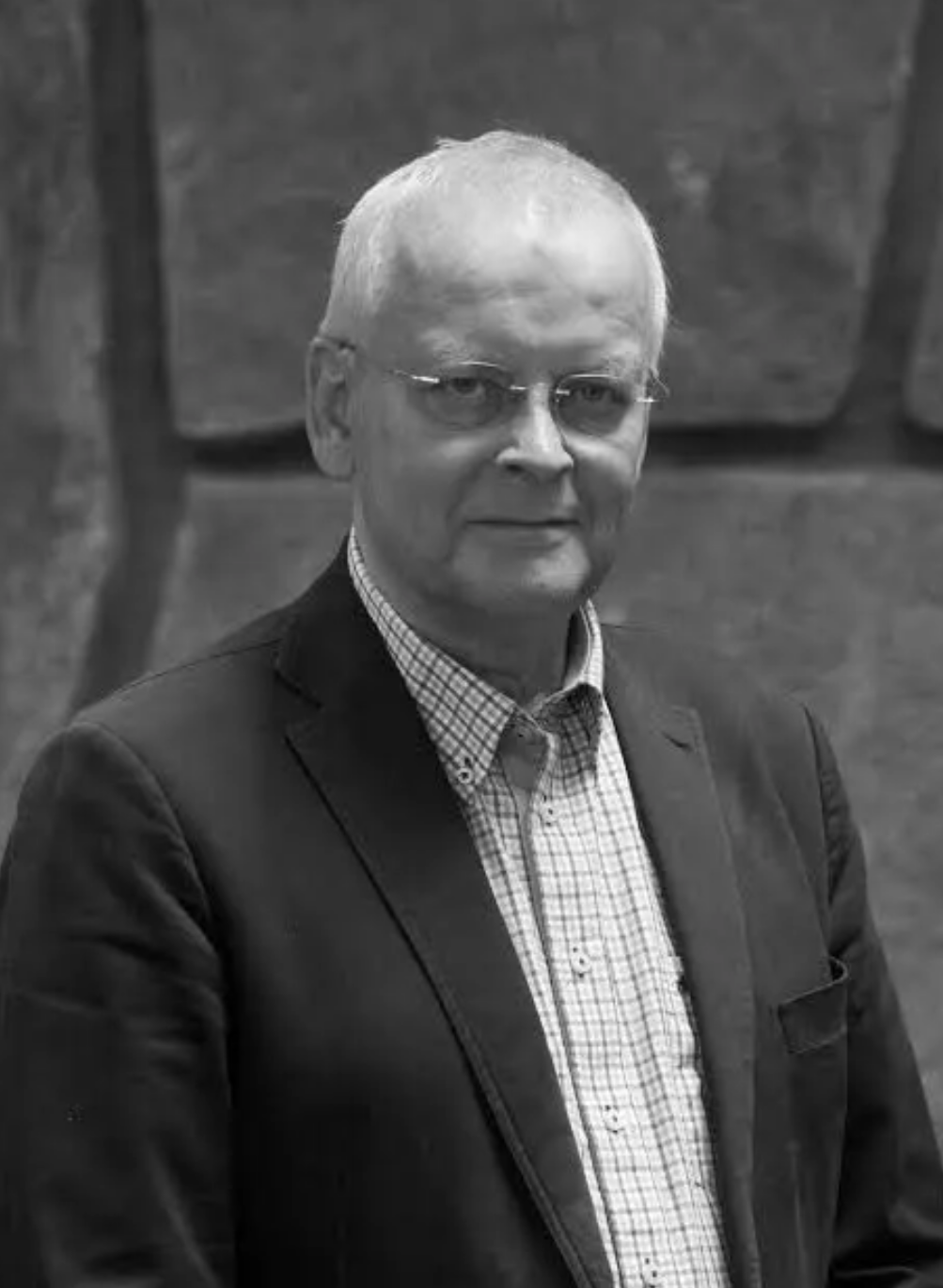 En äldre man med kort vitt hår och glasögon, i en rutig skjorta och mörk kavaj, står framför en stenmur.