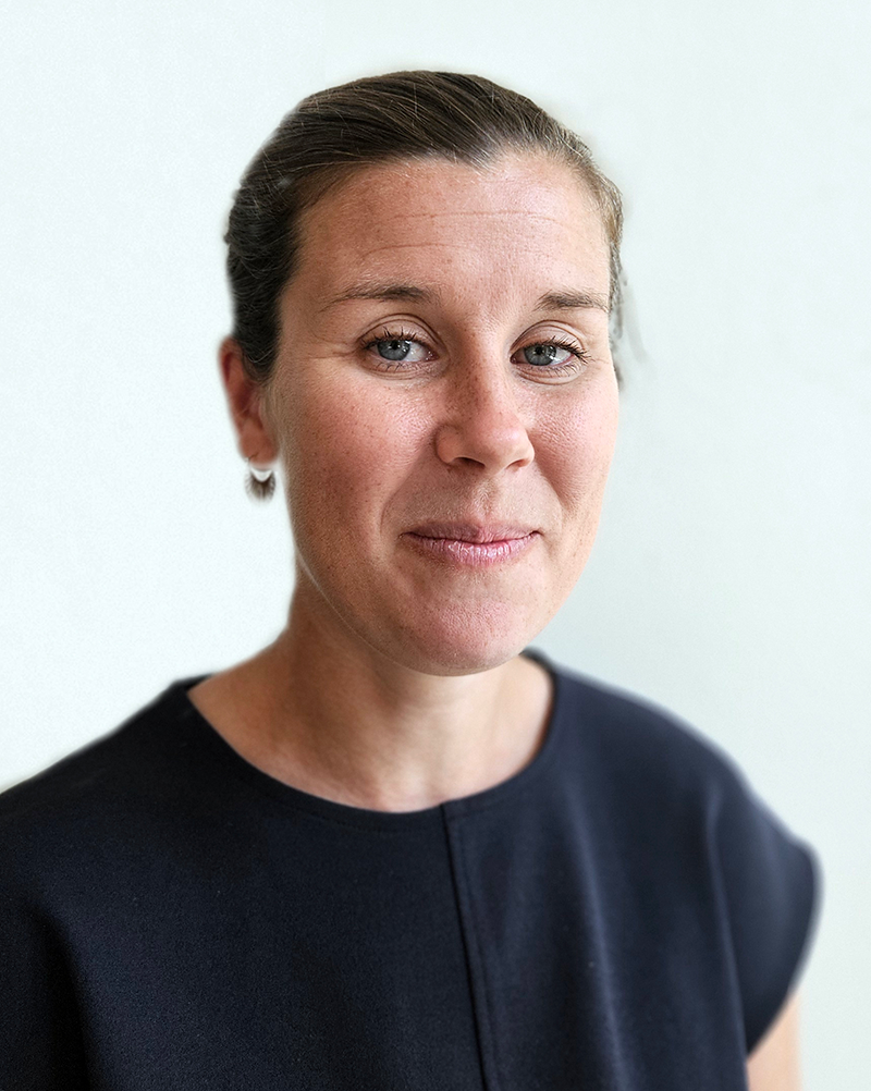 En kvinna med ljus hud och brunt hår uppsatt har en mörk tröja och står framför en ljus bakgrund, vänd mot kameran och ler.