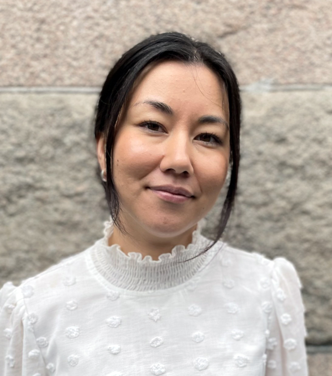 En kvinna med mörkt hår uppsatt har på sig en vit blus och står framför en mur av sten.