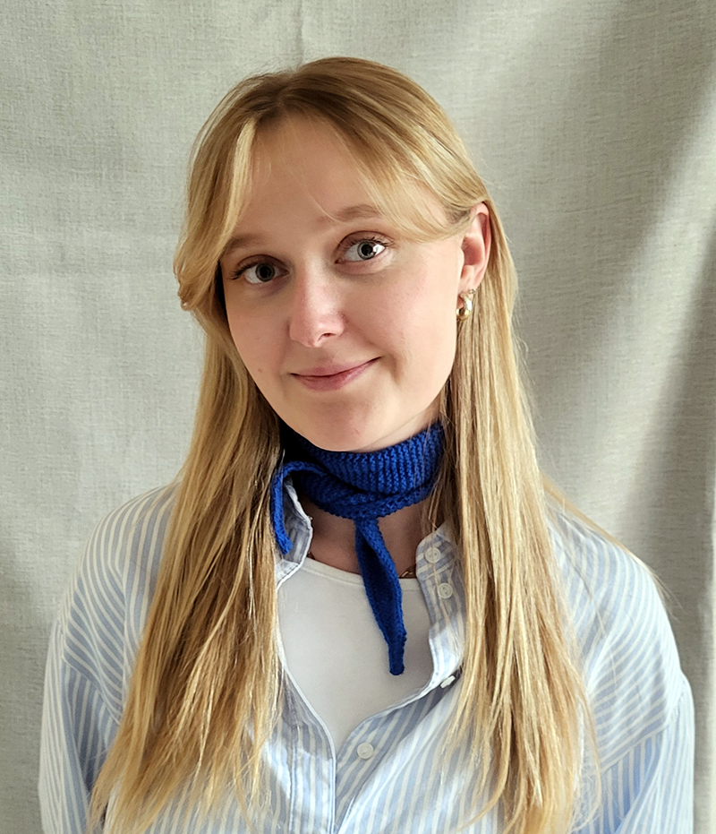 En kvinna med långt blont hår har en blå scarf och en ljusblårandig skjorta. Hon står framför en neutral tygbakgrund.