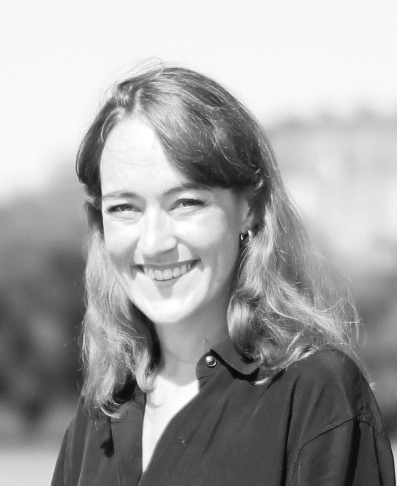 En kvinna med axellångt hår och en skjorta ler utomhus med suddiga träd och byggnader i bakgrunden.