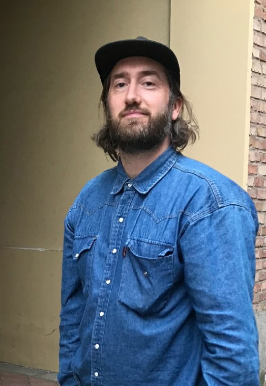 En man med långt hår och skägg, iförd svart keps och blå jeansskjorta, står framför en beige vägg och en hörna av tegel.