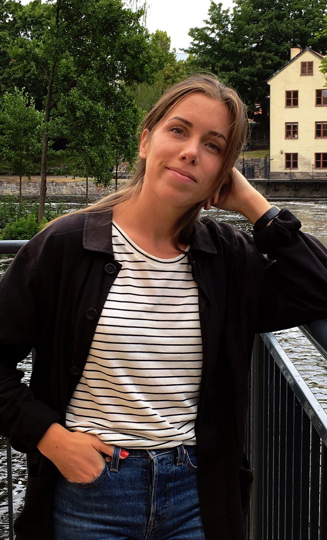 En kvinna i randig tröja och svart jacka lutar sig mot ett räcke vid en flod, med träd och en gul byggnad i bakgrunden.