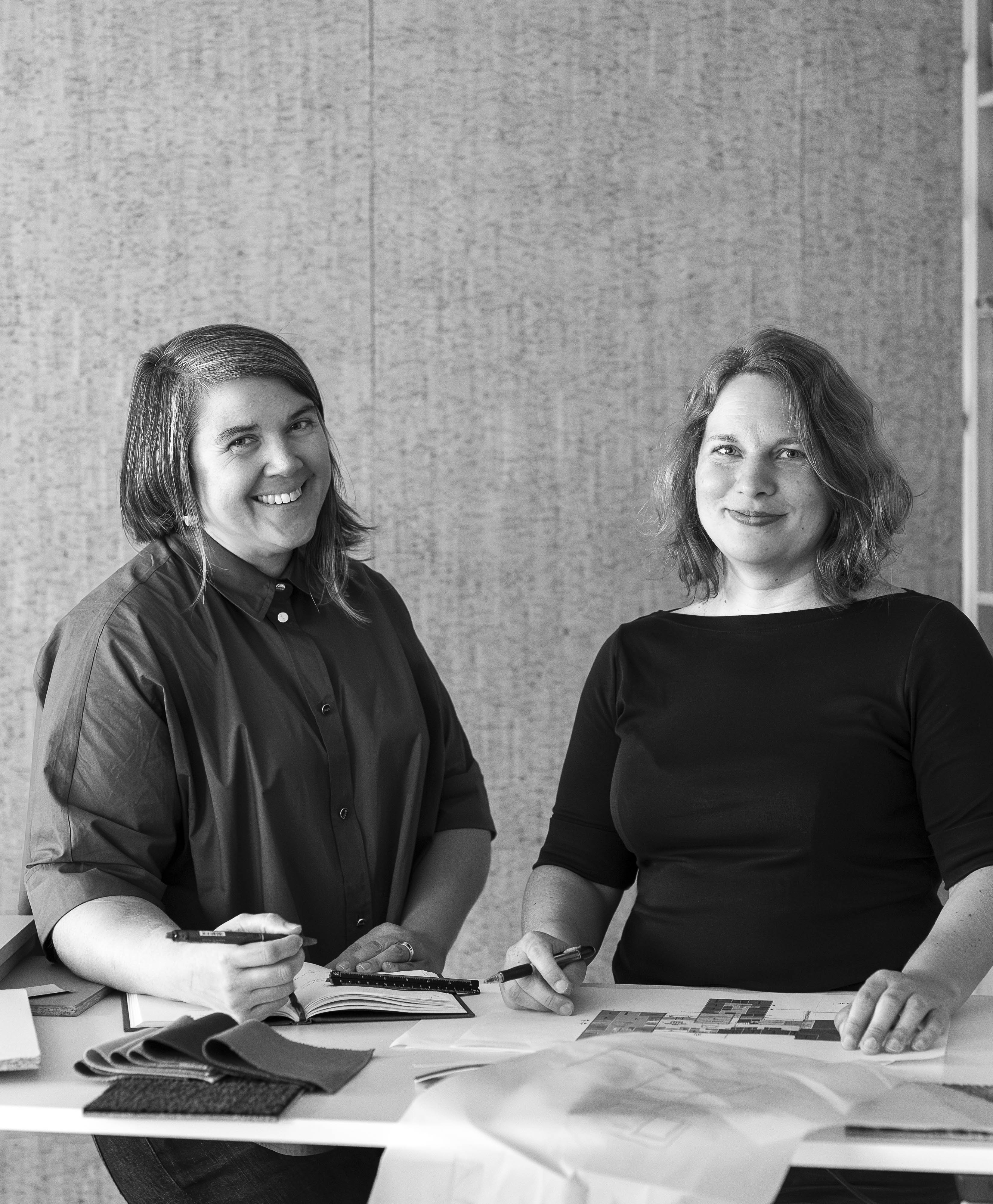 Två kvinnor sitter vid ett bord med papper, tygprover och anteckningsböcker, tittar mot kameran och ler på ett kontor.