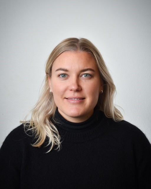 En kvinna med ljust blont hår i svart polotröja står framför en vit bakgrund och ser rakt in i kameran utan uttryck.