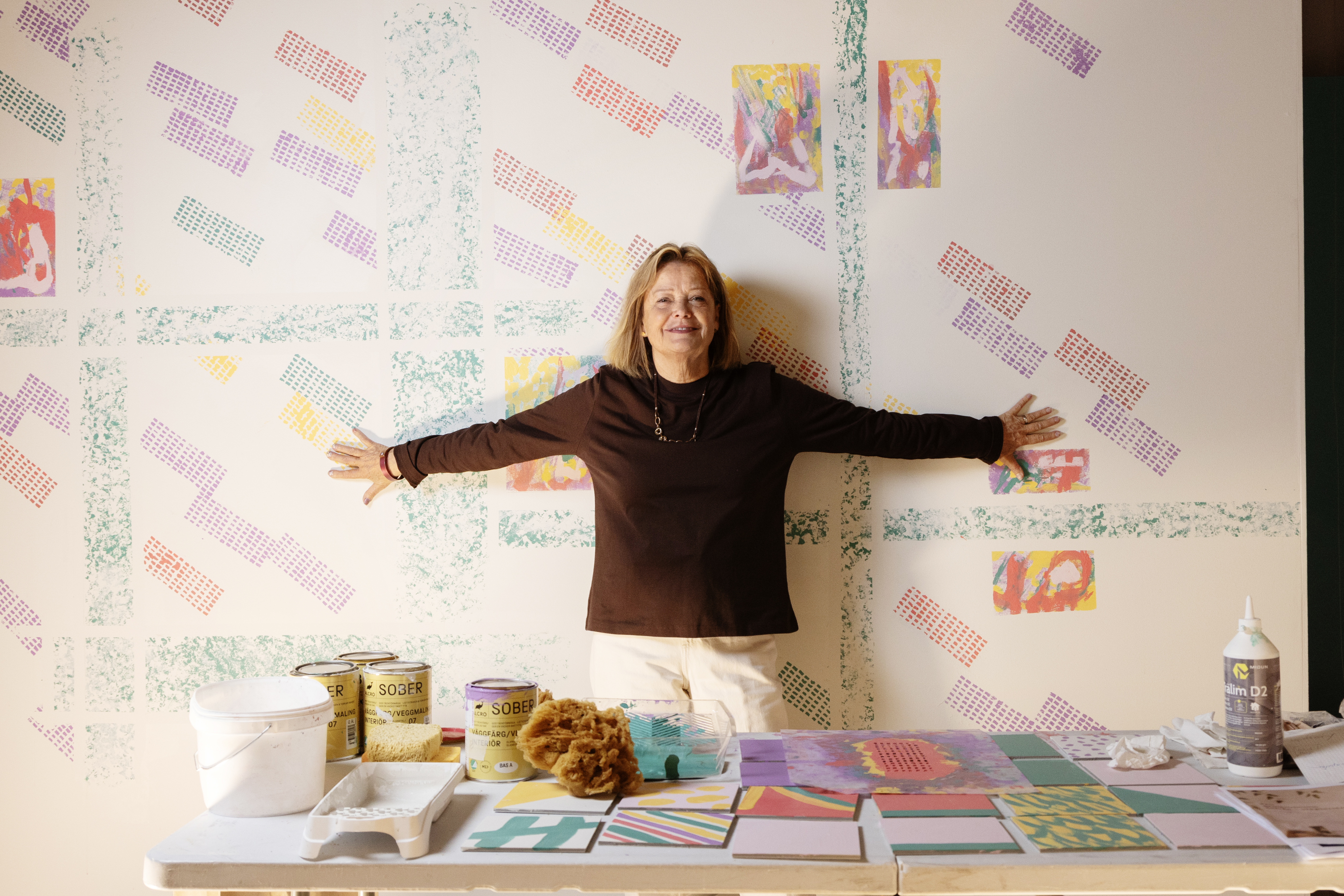 En kvinna står med utsträckta armar framför en dekorerad vägg. Framför henne står burkar med färg, svampar och material.