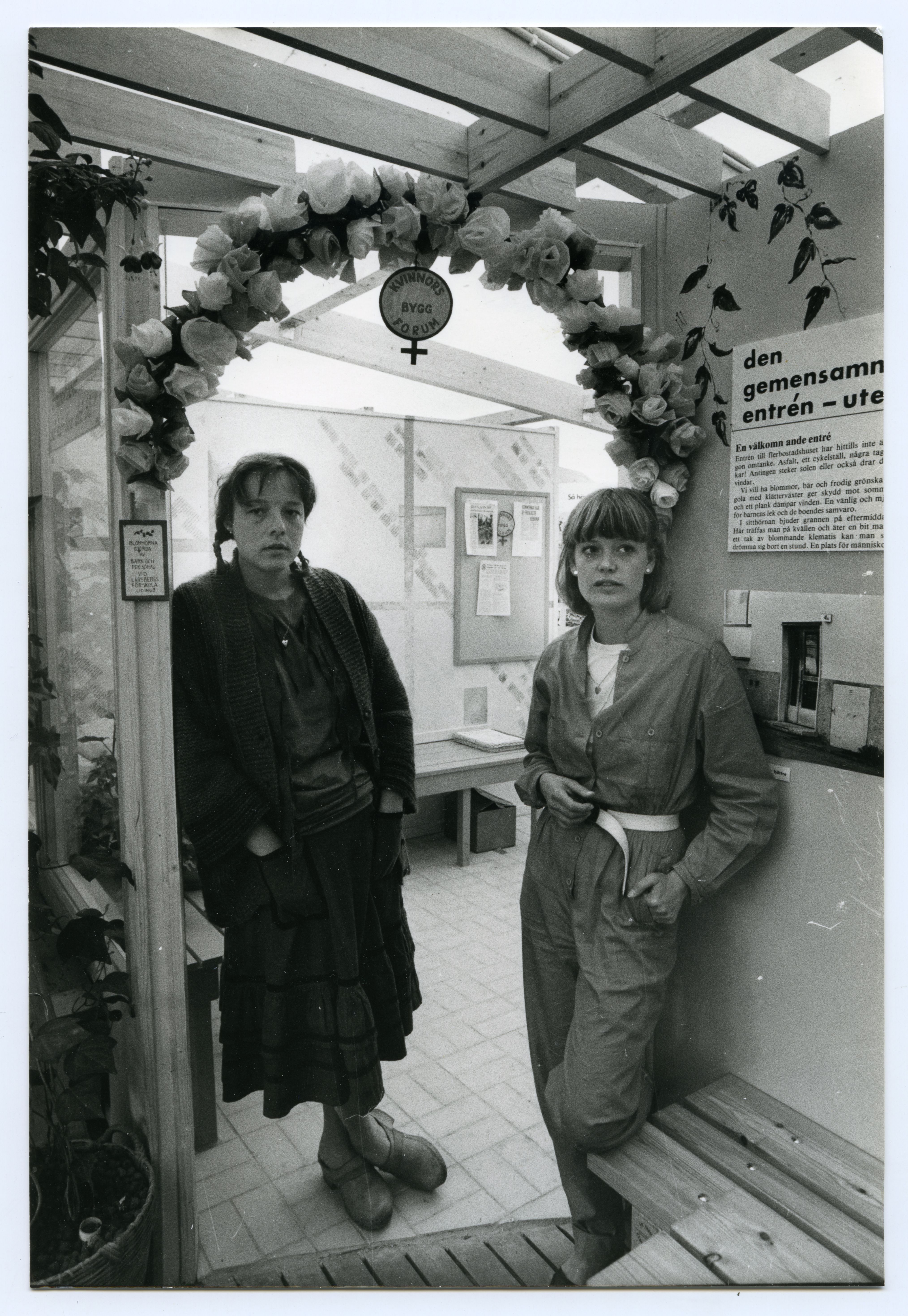 Två kvinnor står under en blomsterbåge i ett rum med informationsaffischer och bänkar. Ena kvinnan har händerna i fickorna; båda tittar mot kameran.