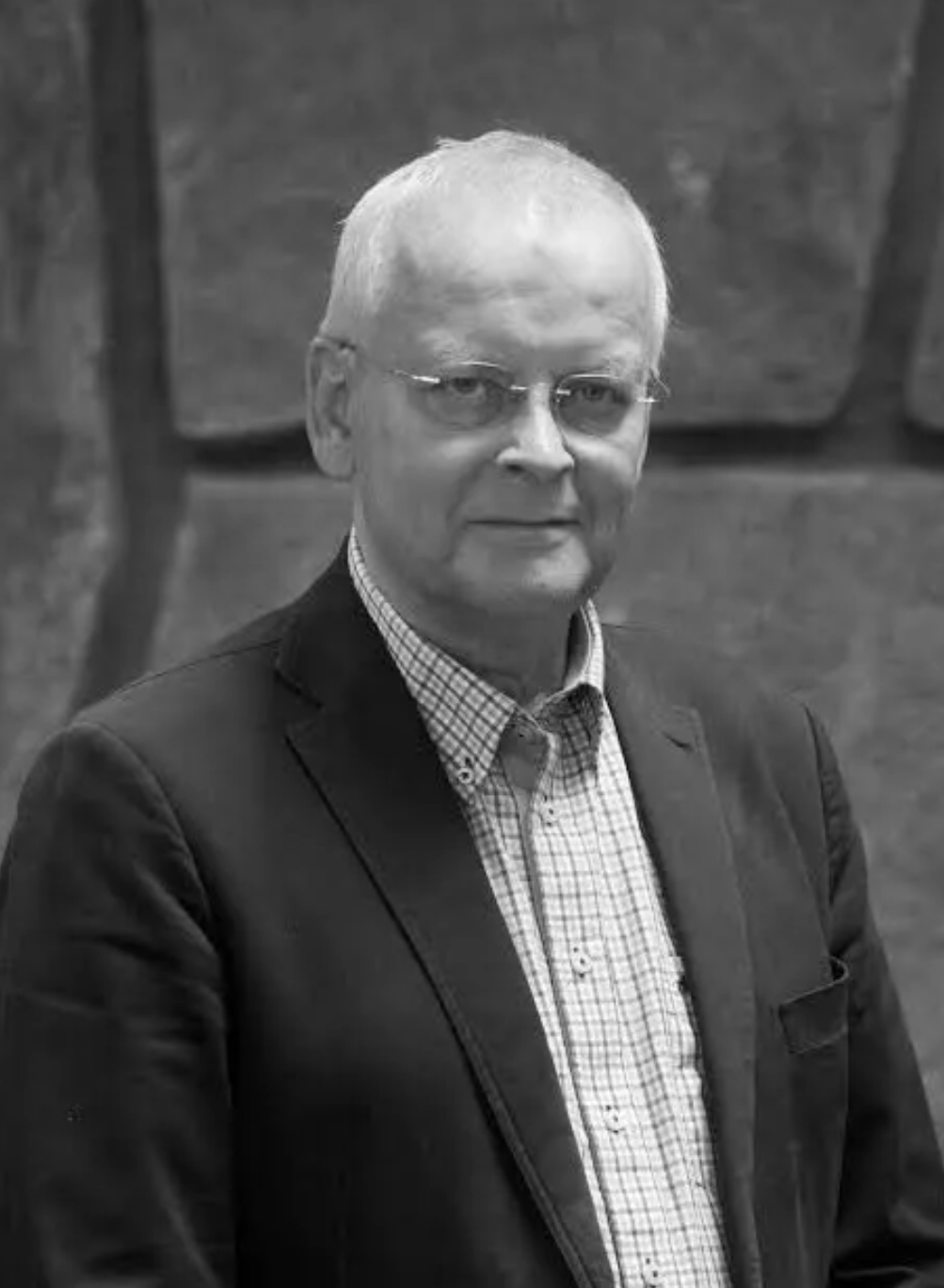 En äldre man med kort vitt hår, glasögon och en kavaj över en rutig skjorta står framför en stenmur och tittar mot kameran.