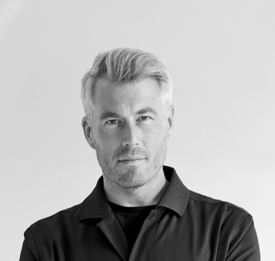 En man med ljust hår i knappad mörk jacka står med korsade armar i en hall med valv. Bilden är i svartvitt.