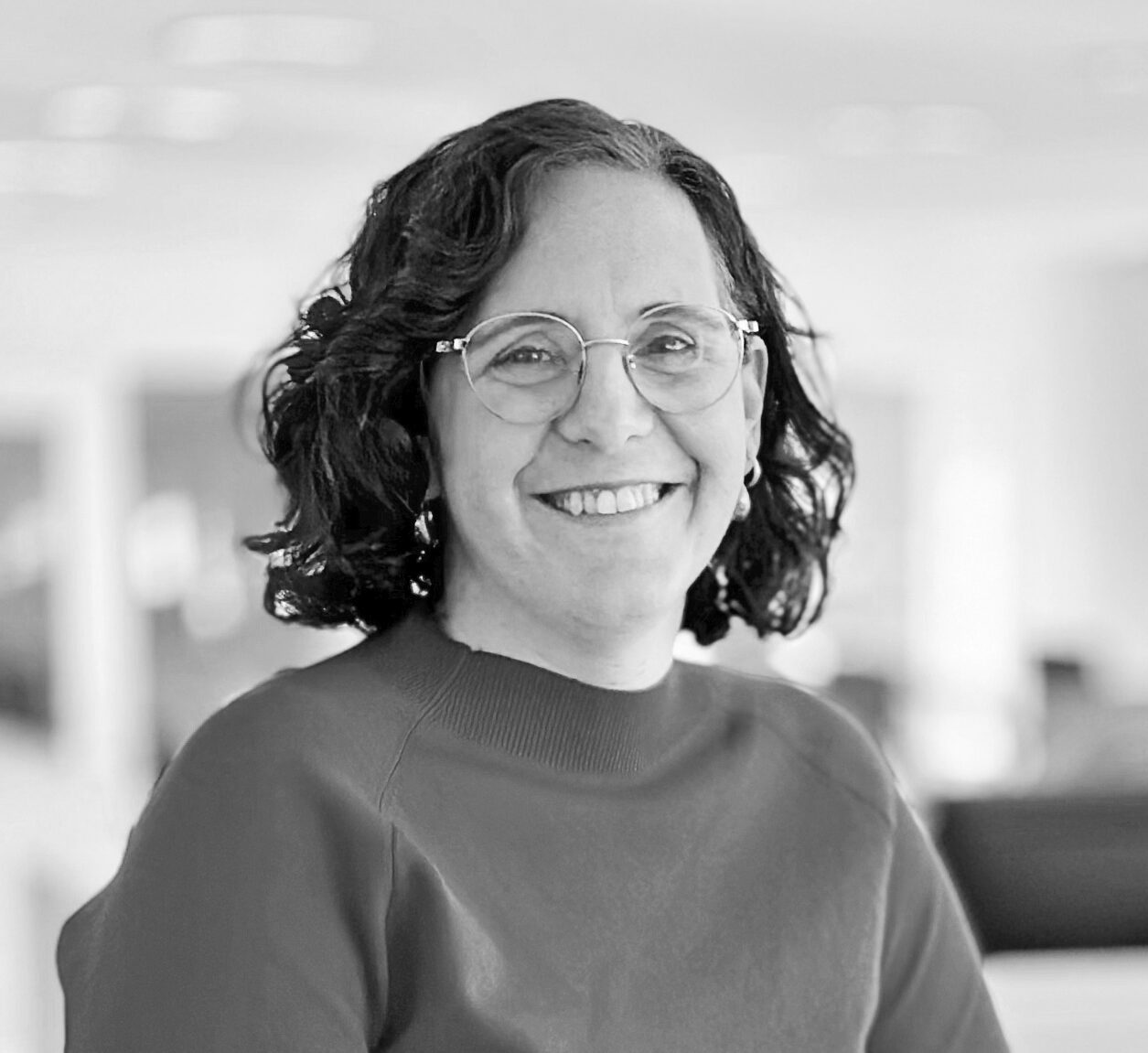 En kvinna med lockigt hår och glasögon står vid ett skrivbord i ett kontor. Hon har långärmad tröja och byxor.