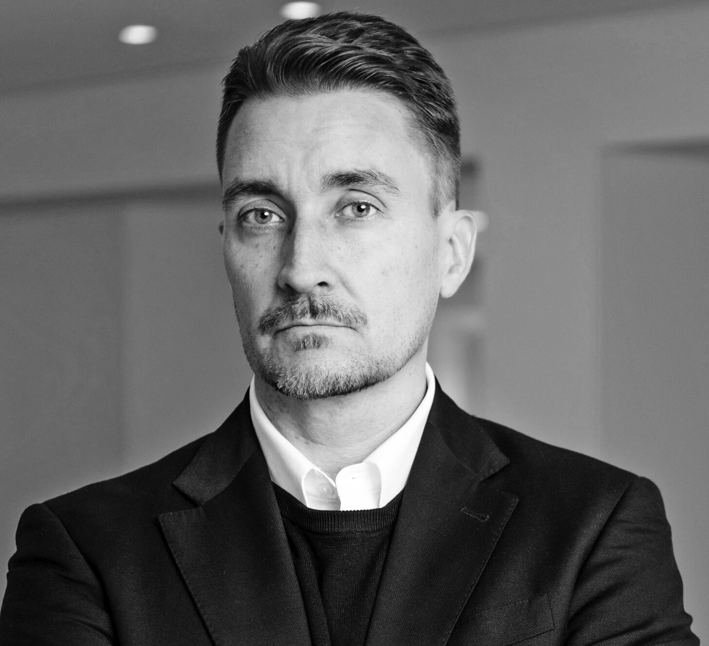 En man i kostym står med armarna i kors i ett kontor och tittar in i kameran. Bilden är svartvit.