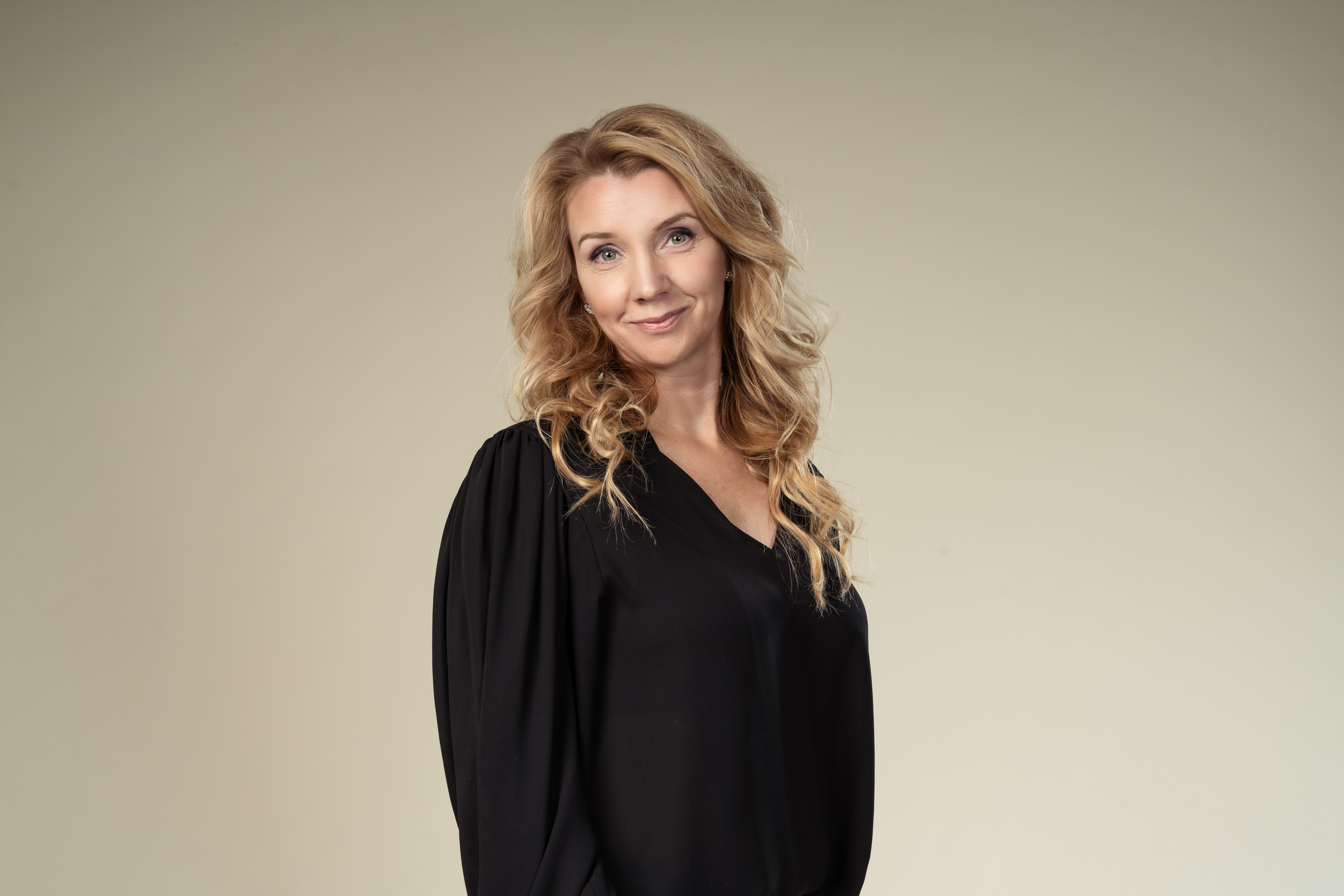 En kvinna med långt, vågigt blont hår och svart blus står mot en beige bakgrund, tittar mot kameran och ler svagt.