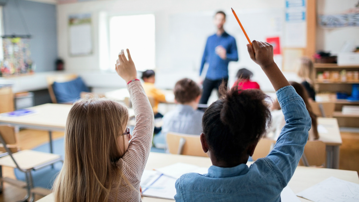 Barn sitter vid bänkar i ett klassrum och räcker upp händerna, medan en lärare står och talar längst fram i rummet.