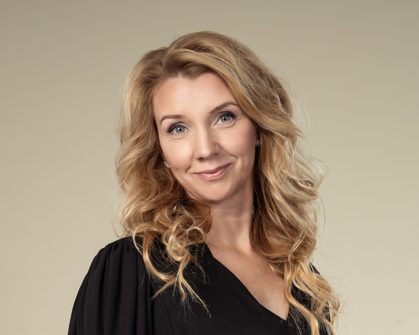 En kvinna med långt, vågigt blont hår och svart blus står mot en beige bakgrund, tittar mot kameran och ler svagt.
