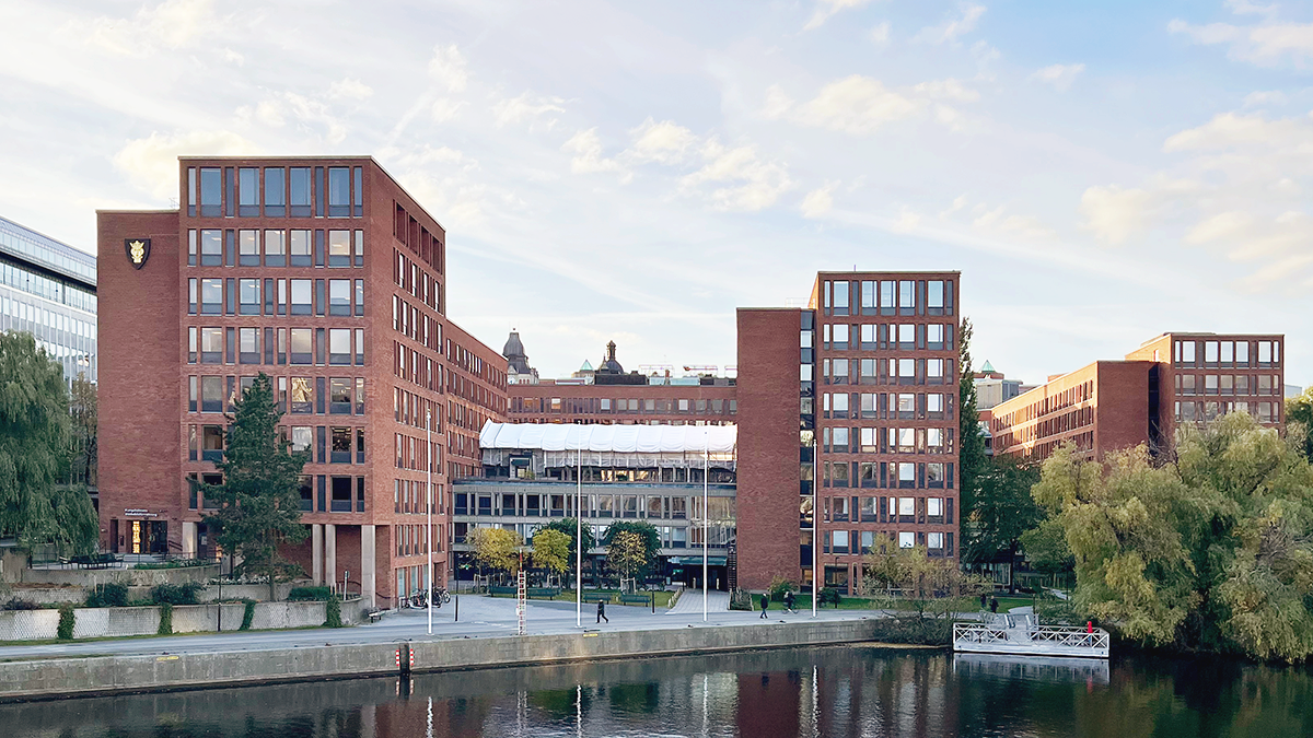 Ett kontorskomplex i rött tegel ligger vid vattnet, med träd och blå himmel i bakgrunden.